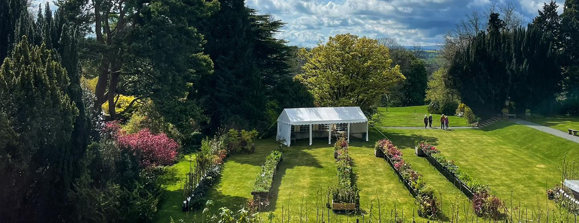 An aerial shot of an event in the gardens with. long tables displaying flowers and plants. A marquee behind and a few people milling around